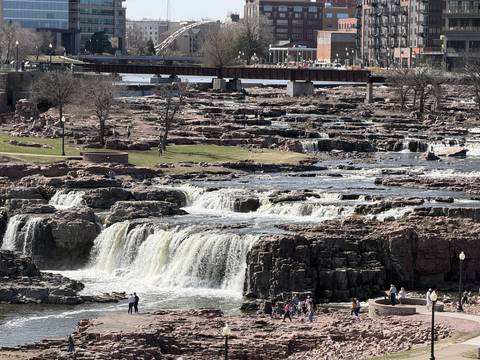 The falls of the Big Sioux River pouring over pink quartzite, with the railroad bridge and downtown Sioux Falls behind